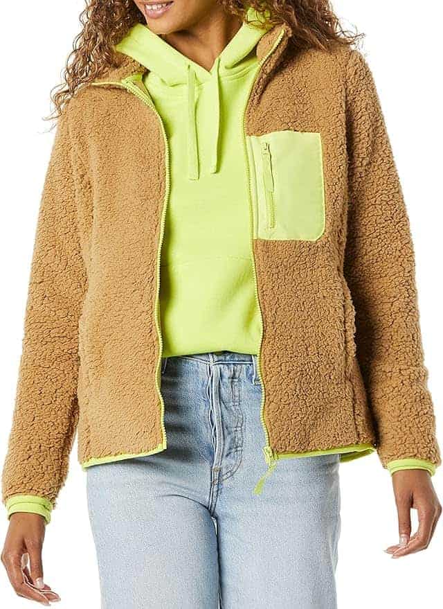 A person wearing a tan fleece jacket with a neon yellow zip pocket over a neon yellow hoodie and light blue jeans.