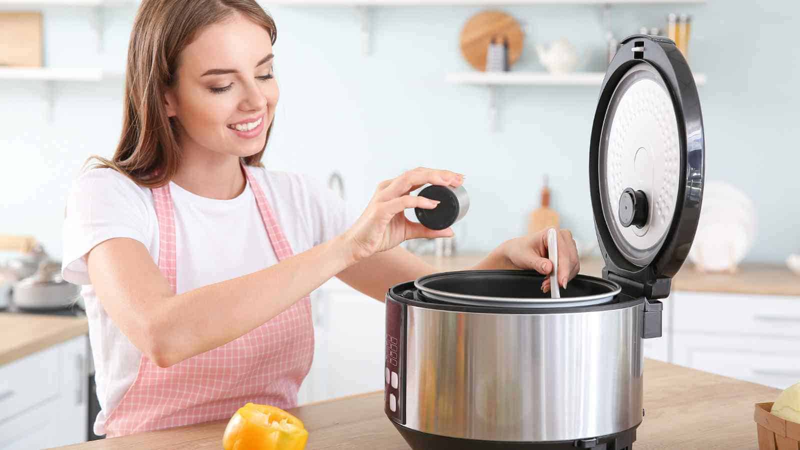 A woman in a pink apron places an ingredient into an open electric pressure cooker on a kitchen counter.