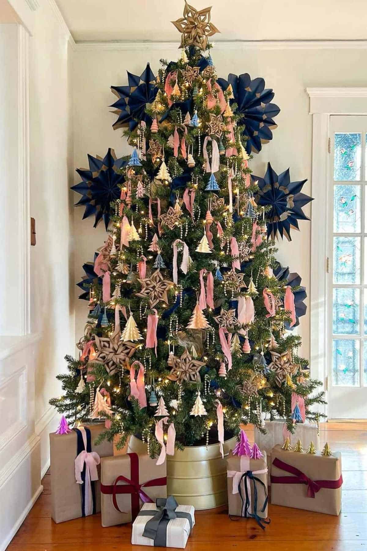 A decorated Christmas tree with gold, pink, and metallic ornaments stands in a living room with wrapped gifts underneath and blue star decorations on the wall behind it.