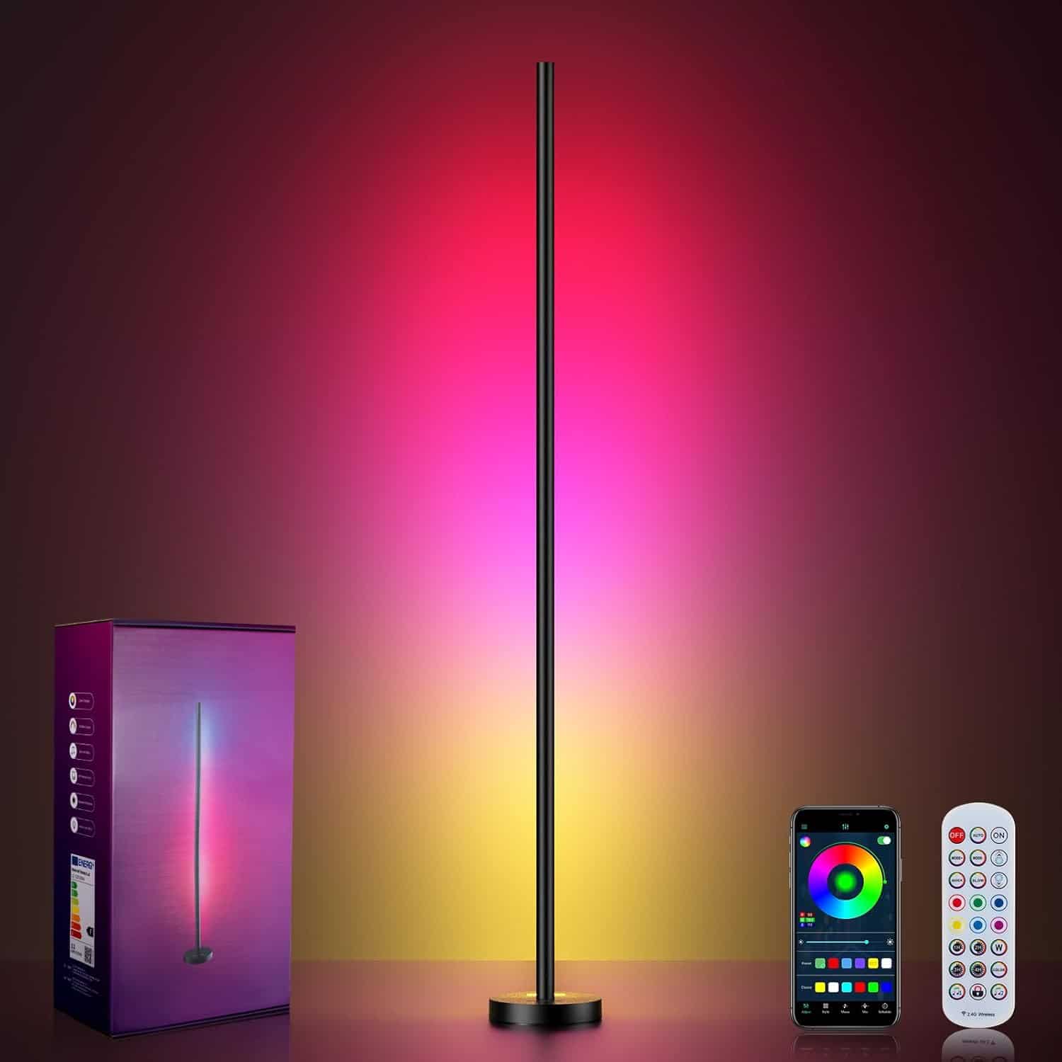A color-changing LED floor lamp stands lit in front of a wall, with a product box, smartphone displaying a color wheel app, and two remote controls beside it.