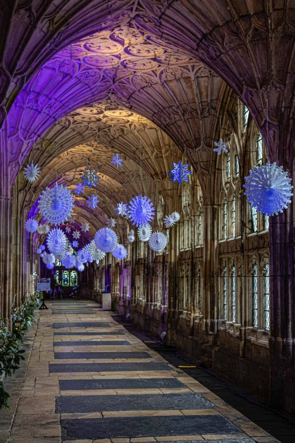 A stone hallway with vaulted ceilings is decorated with hanging white snowflakes and illuminated by purple lighting.