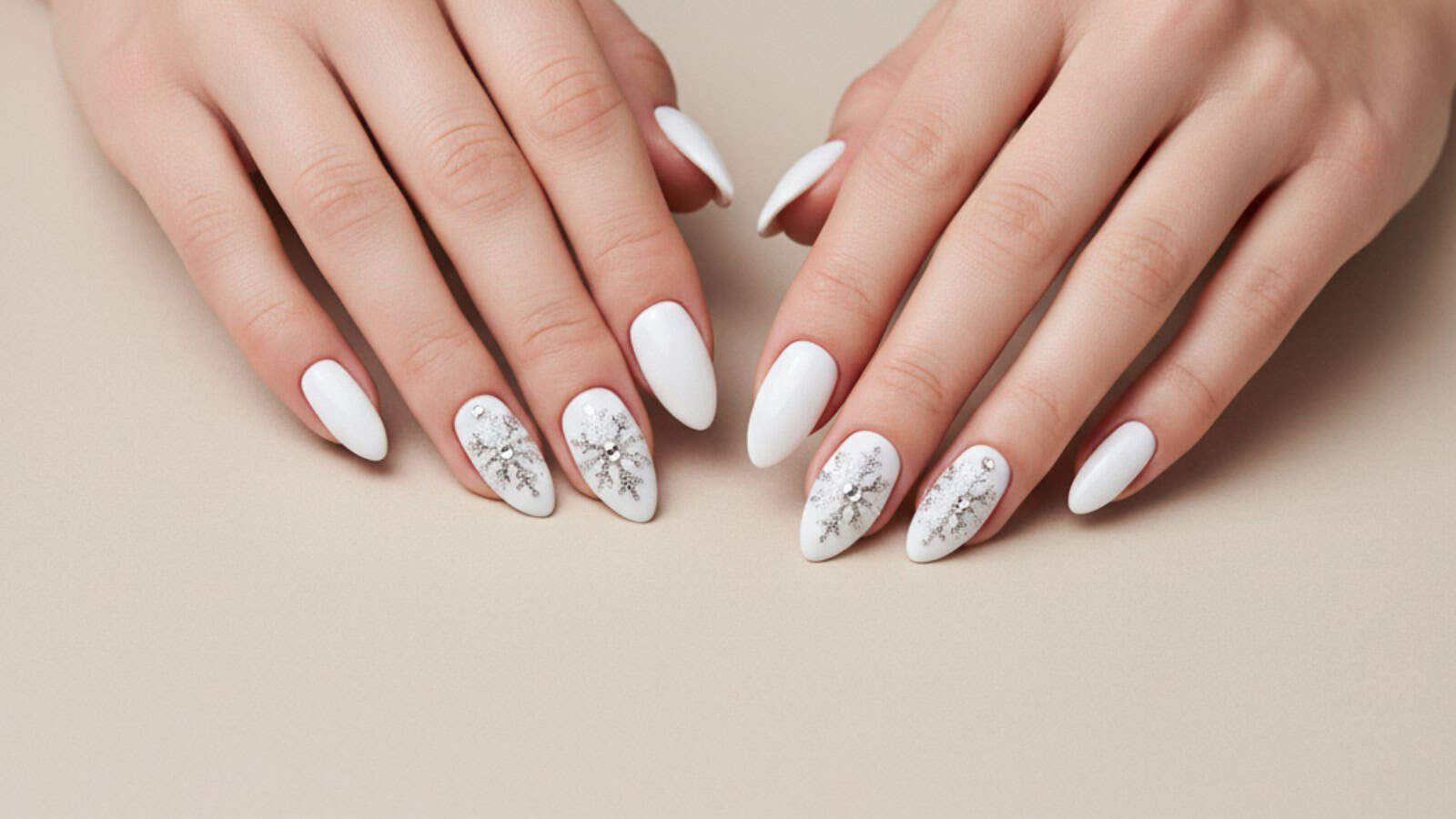 Hands with white, almond-shaped nails. Four nails feature silver snowflakes designs while the rest are plain white. The hands rest on a light beige surface.