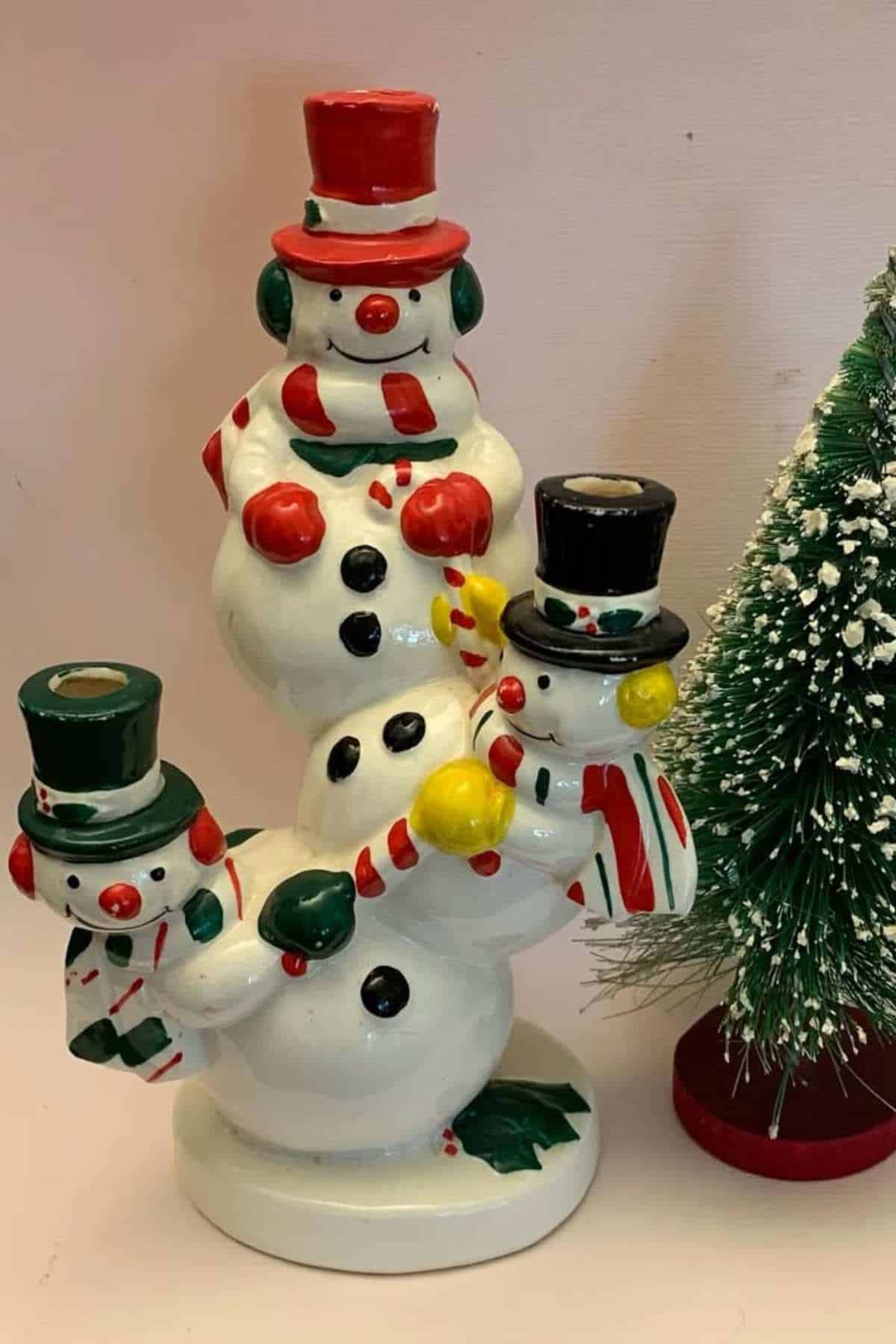 Three ceramic snowmen with colorful hats and scarves are connected in a spiral, next to a small artificial Christmas tree with snow on its branches.