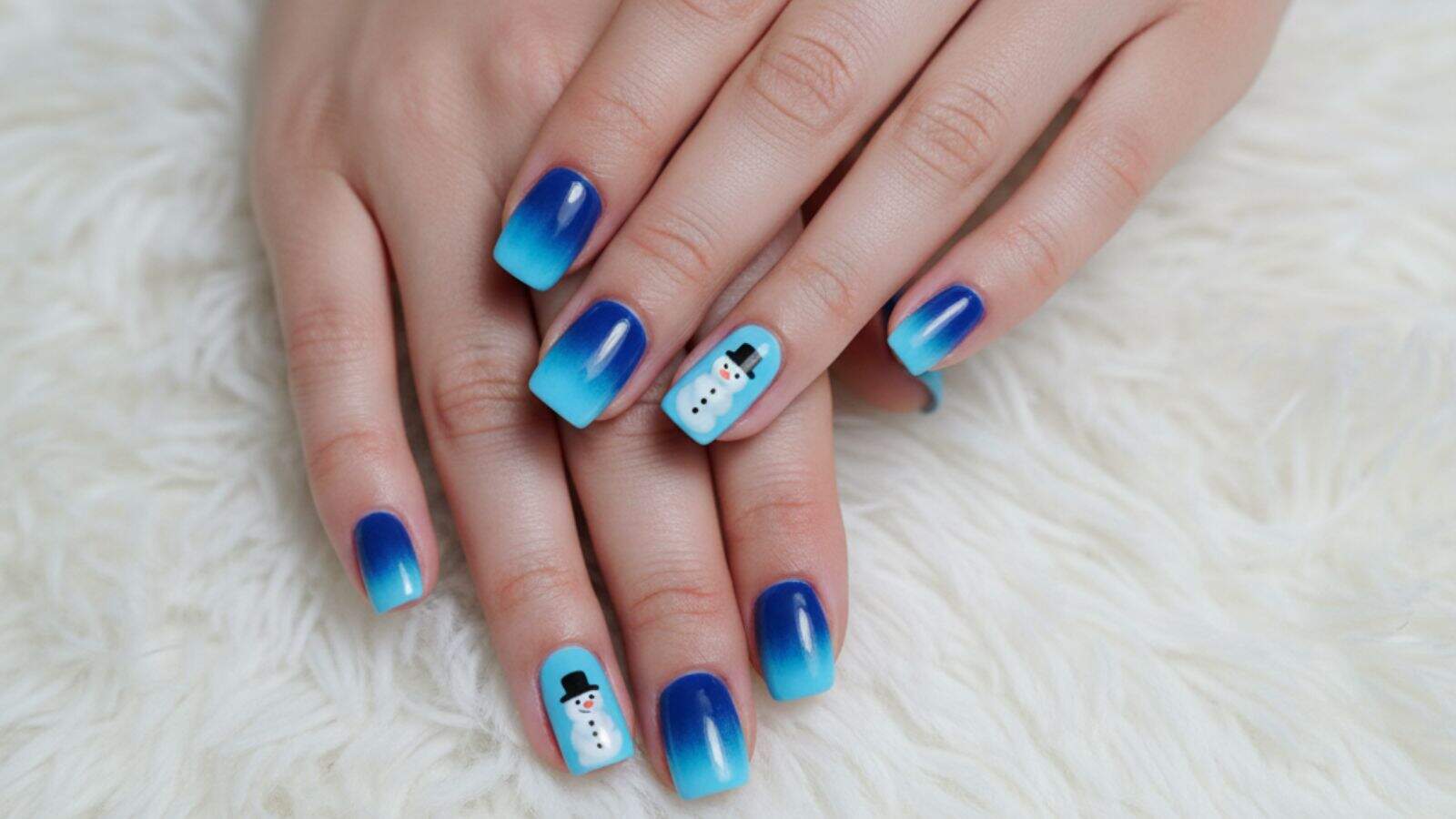 Hands with blue gradient manicure, featuring snowman designs on two accent nails, resting on a white textured surface.