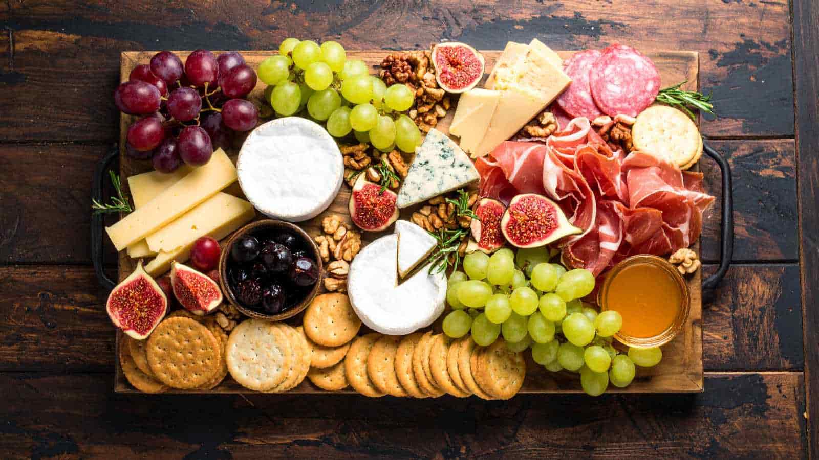 A wooden board with assorted cheeses, crackers, grapes, figs, olives, walnuts, honey, and cured meats arranged neatly.