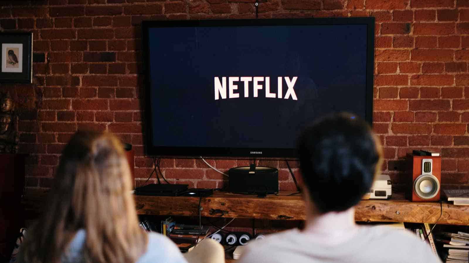 Two people sit on a couch facing a television displaying the Netflix logo in a living room with a brick wall.