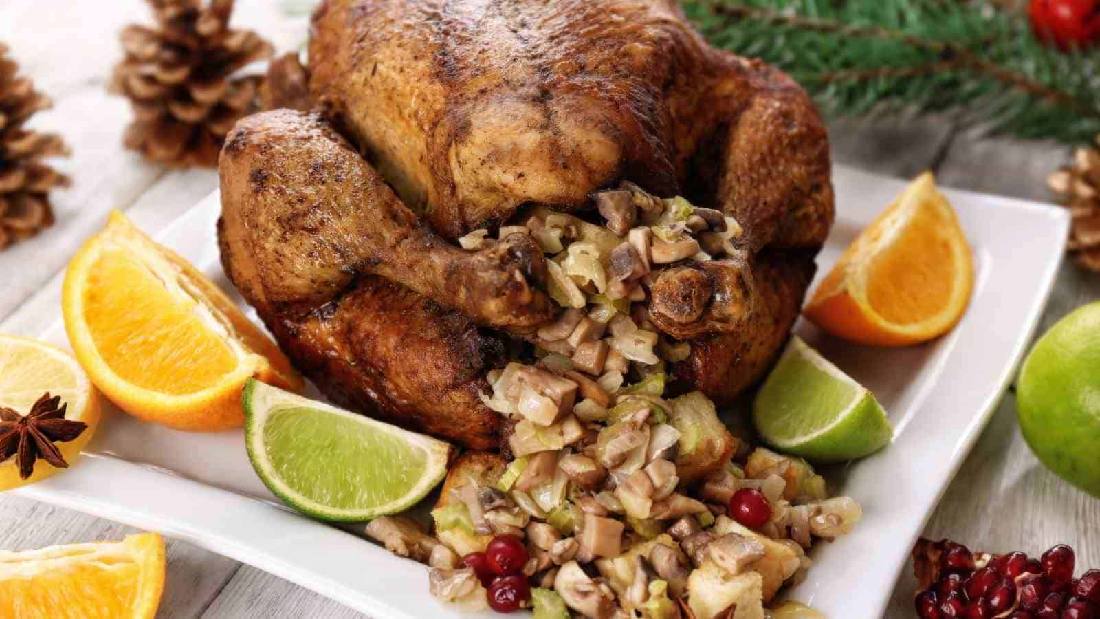 Roast whole chicken stuffed with vegetables and mushrooms, garnished with orange and lime wedges on a white plate. Pinecones and greenery in the background.