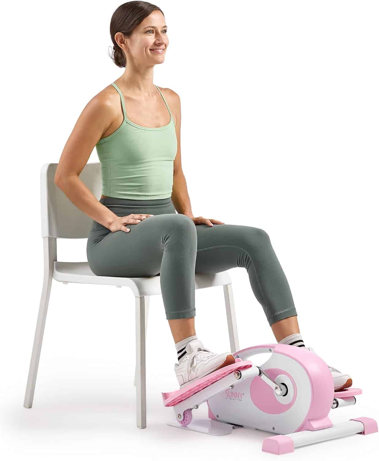 Woman sitting on a chair using a pink and white pedal exerciser, wearing a green tank top and gray leggings, with a relaxed and upright posture.