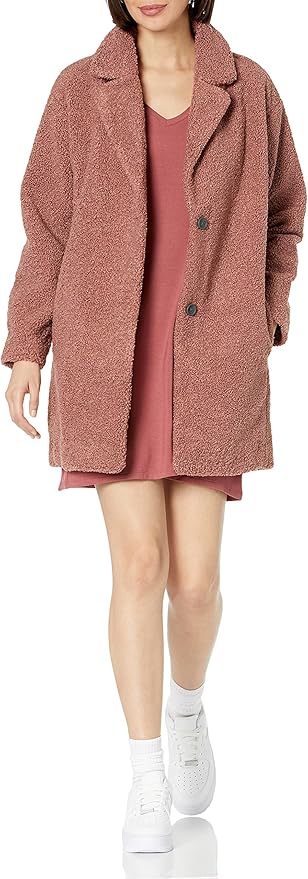 A person wearing a textured pinkish-brown coat over a pink dress, white socks, and white sneakers, walking forward against a plain background.