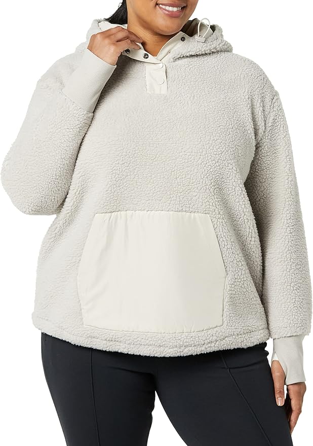 Person wearing a light-colored sherpa hoodie with a large front pocket and black pants, standing against a plain background.