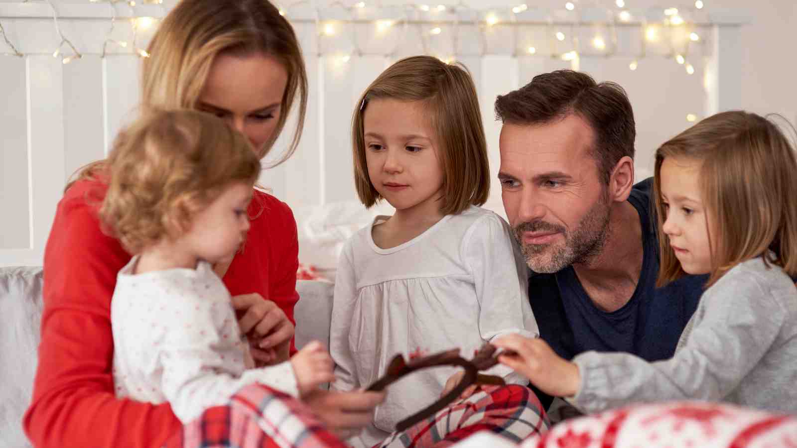 A family with two adults and three young children sit together on a bed, looking at an object, with string lights in the background.