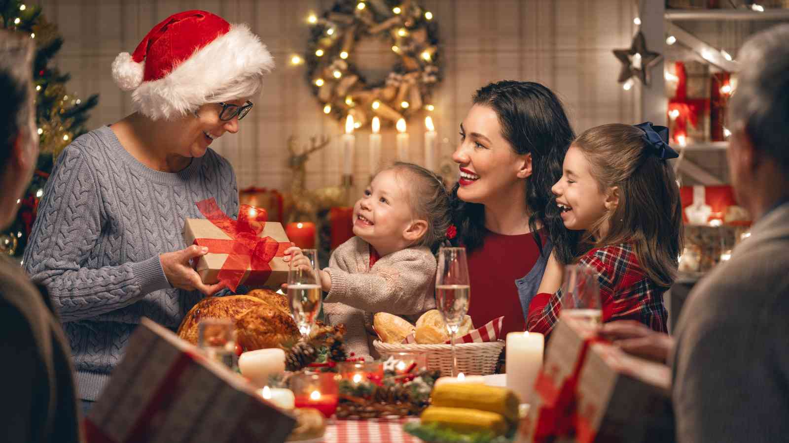 A family gathers around a table with food and candles, exchanging gifts and smiling, with festive holiday decorations in the background.