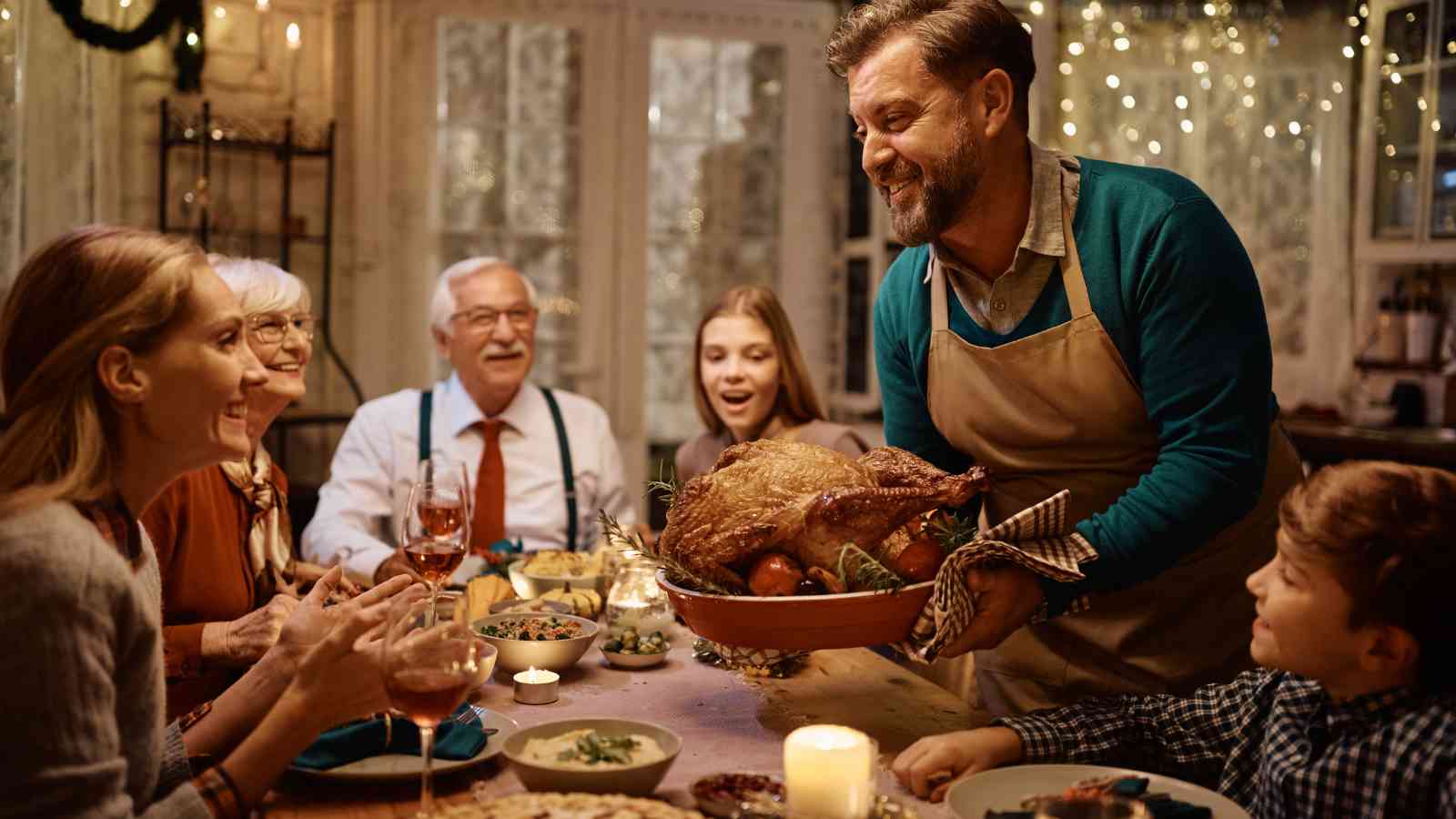 A man wearing an apron presents a roast turkey to a group of people seated around a decorated dinner table during a festive meal.