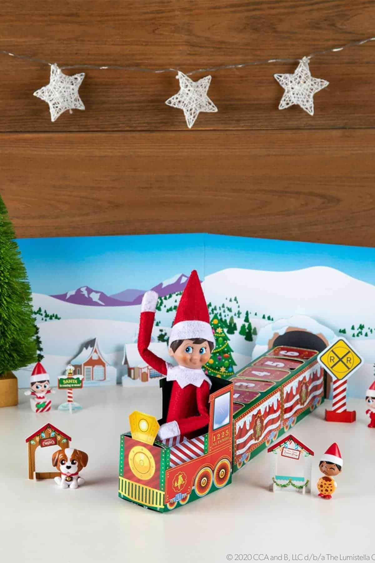 A toy elf in red sits on a colorful paper train with holiday decorations, a small dog figure, and miniature signs; snowy village backdrop and star ornaments overhead.