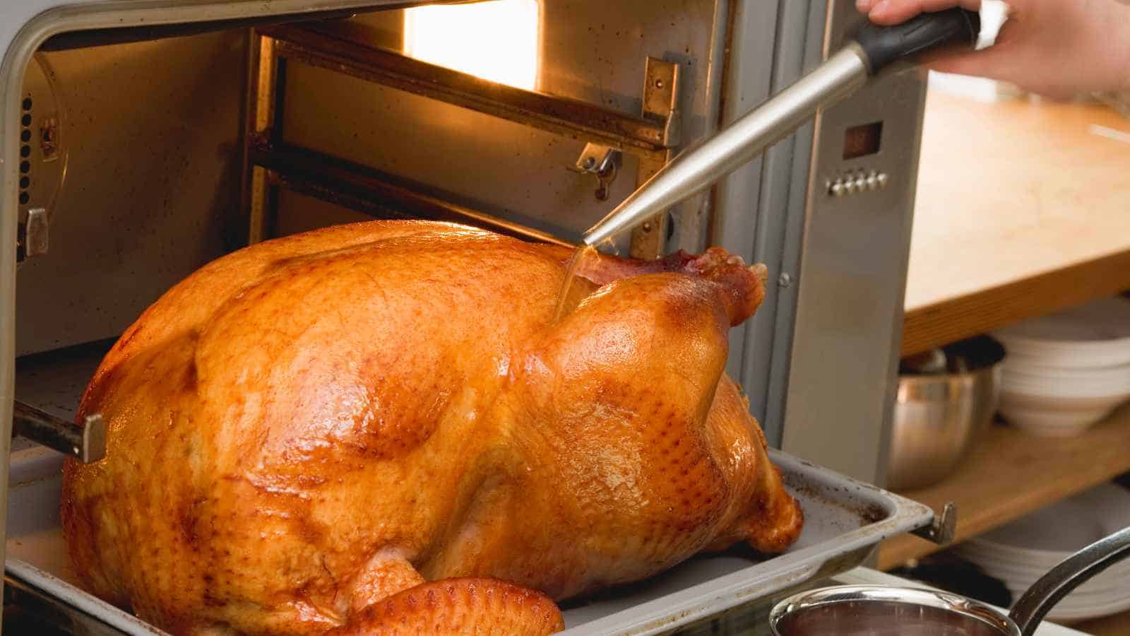 A person uses a baster to moisten a roasted turkey on a tray, with the bird partially inside an oven.
