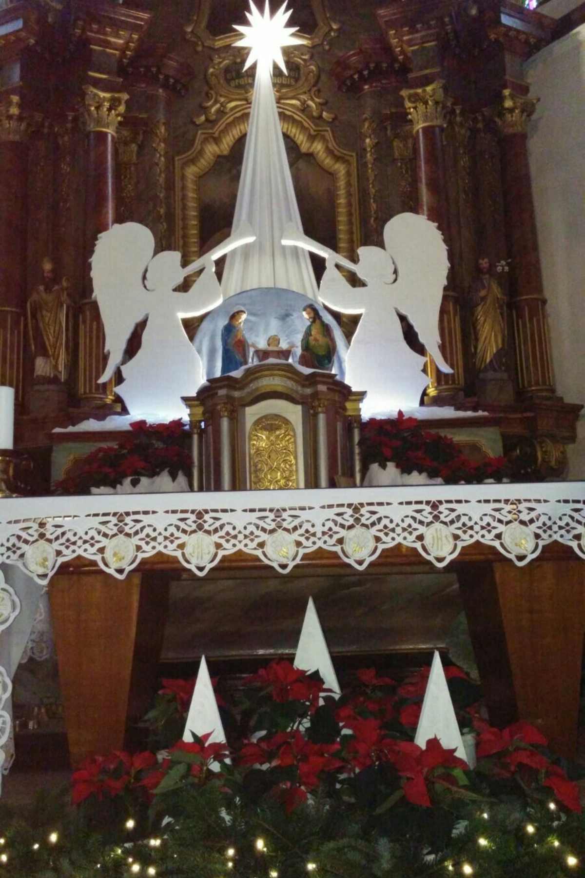 A nativity scene with two angel cutouts, Mary and Joseph figures, a manger, white cloth decorations, red poinsettias, and lights displayed on an ornate altar.