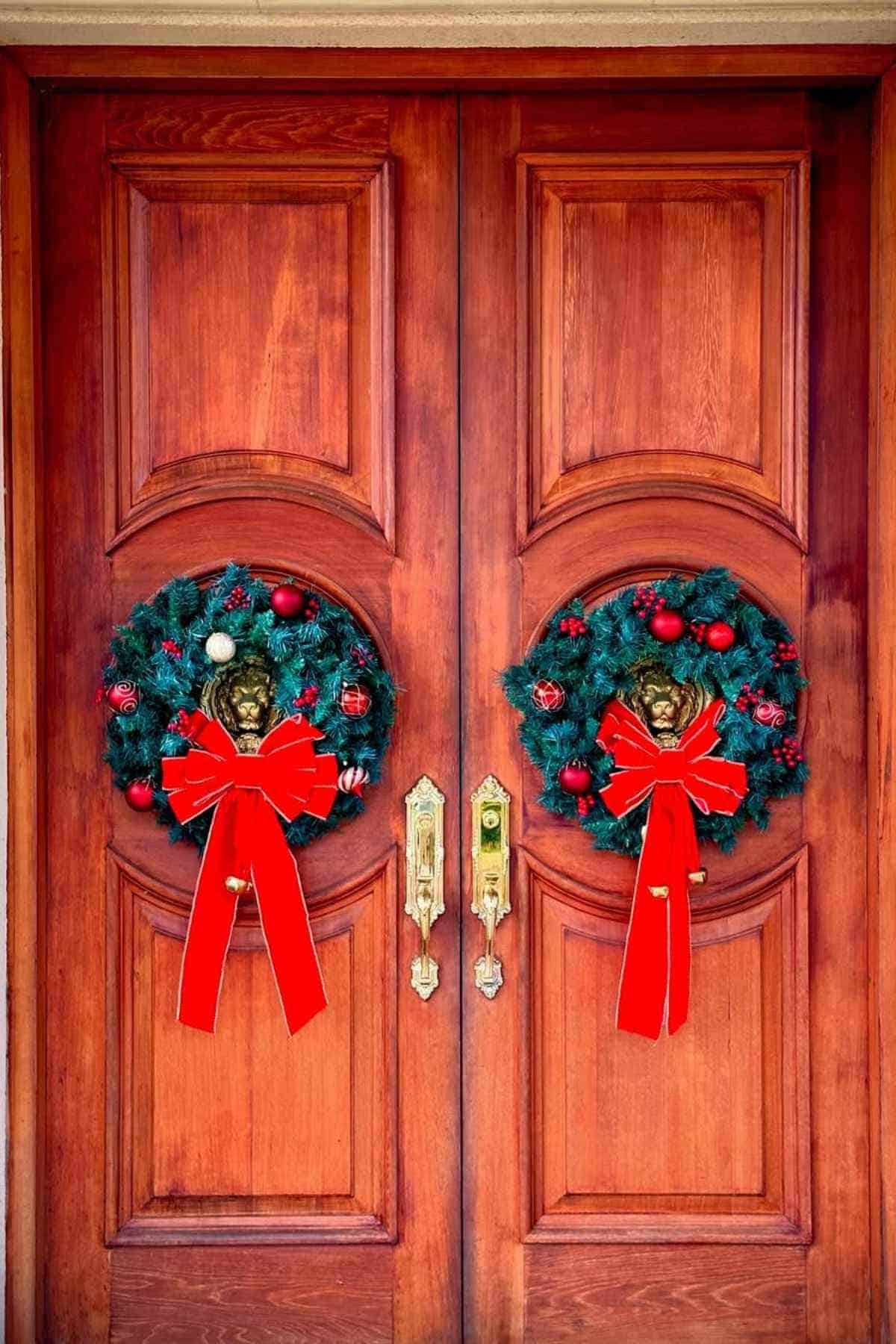 Double wooden doors decorated with two holiday wreaths, each adorned with red ornaments, pinecones, and a large red bow, featuring lion head door knockers.