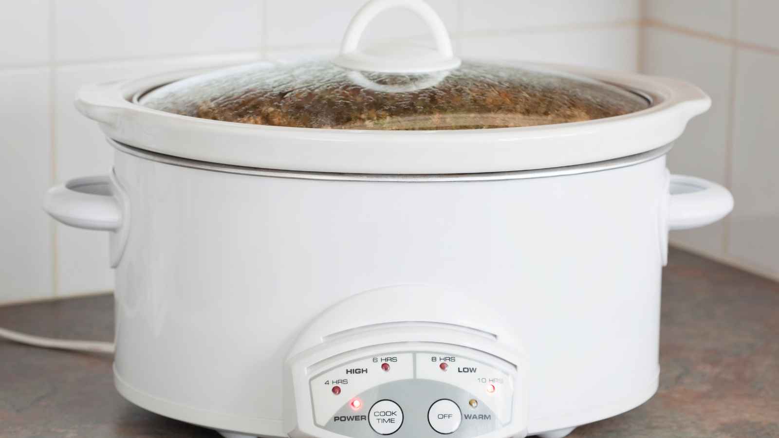 A white slow cooker with a glass lid sits on a kitchen counter, plugged in and turned on, with food cooking inside.