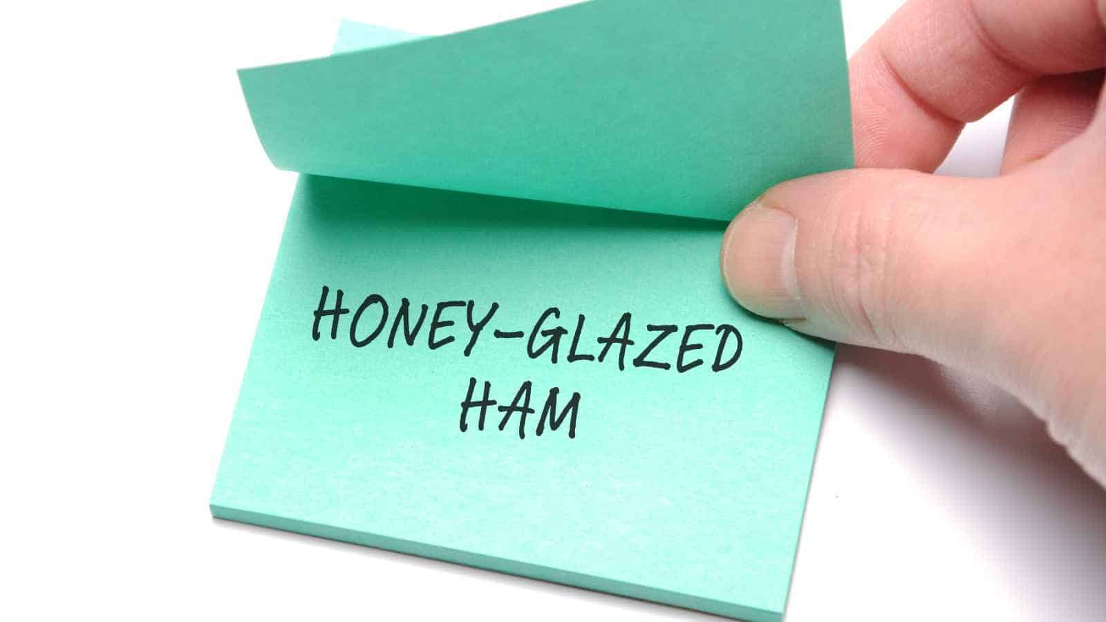 A hand lifts the top sheet of a green sticky note with the words "HONEY-GLAZED HAM" written in black marker.