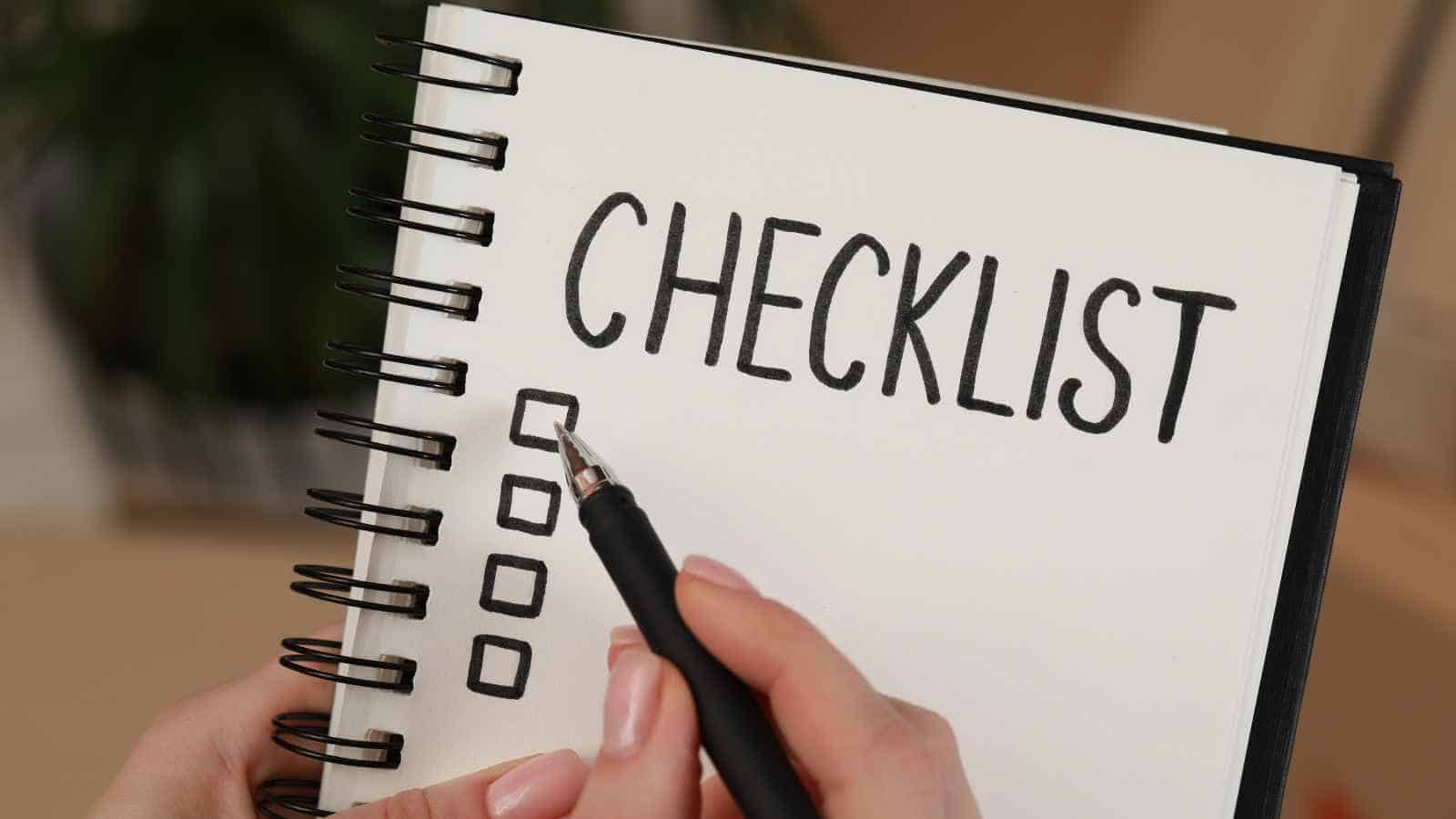 A person holding a pen prepares to write on a spiral notebook with the word "CHECKLIST" written at the top and five empty checkboxes below.