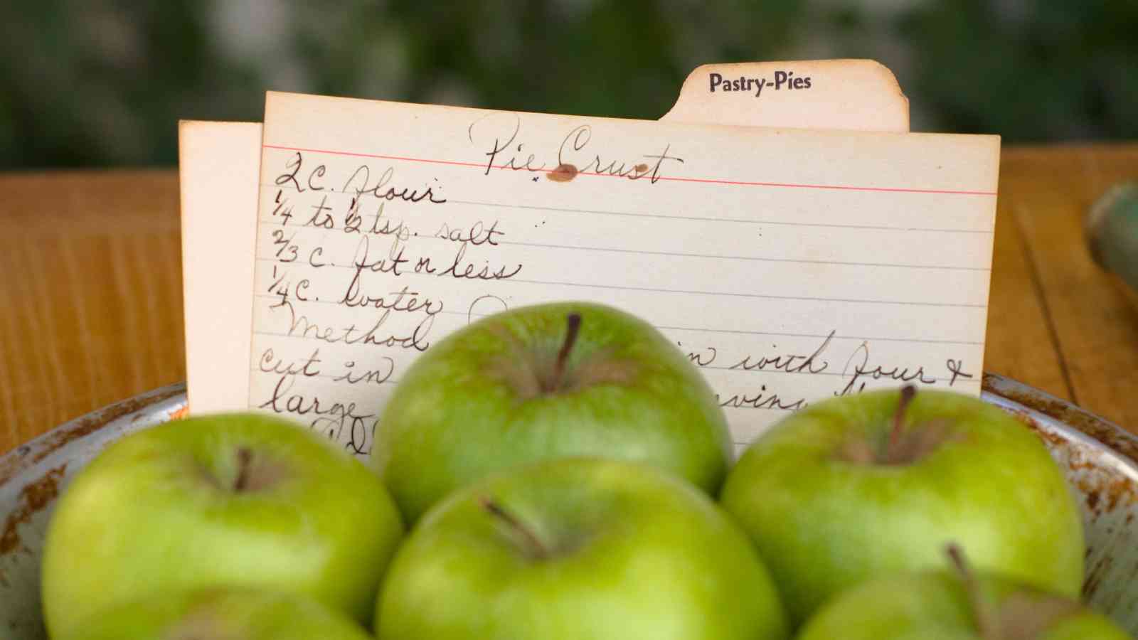 A bowl of green apples sits in front of a handwritten recipe card labeled "Pie Crust" under a "Pastry-Pies" tab.