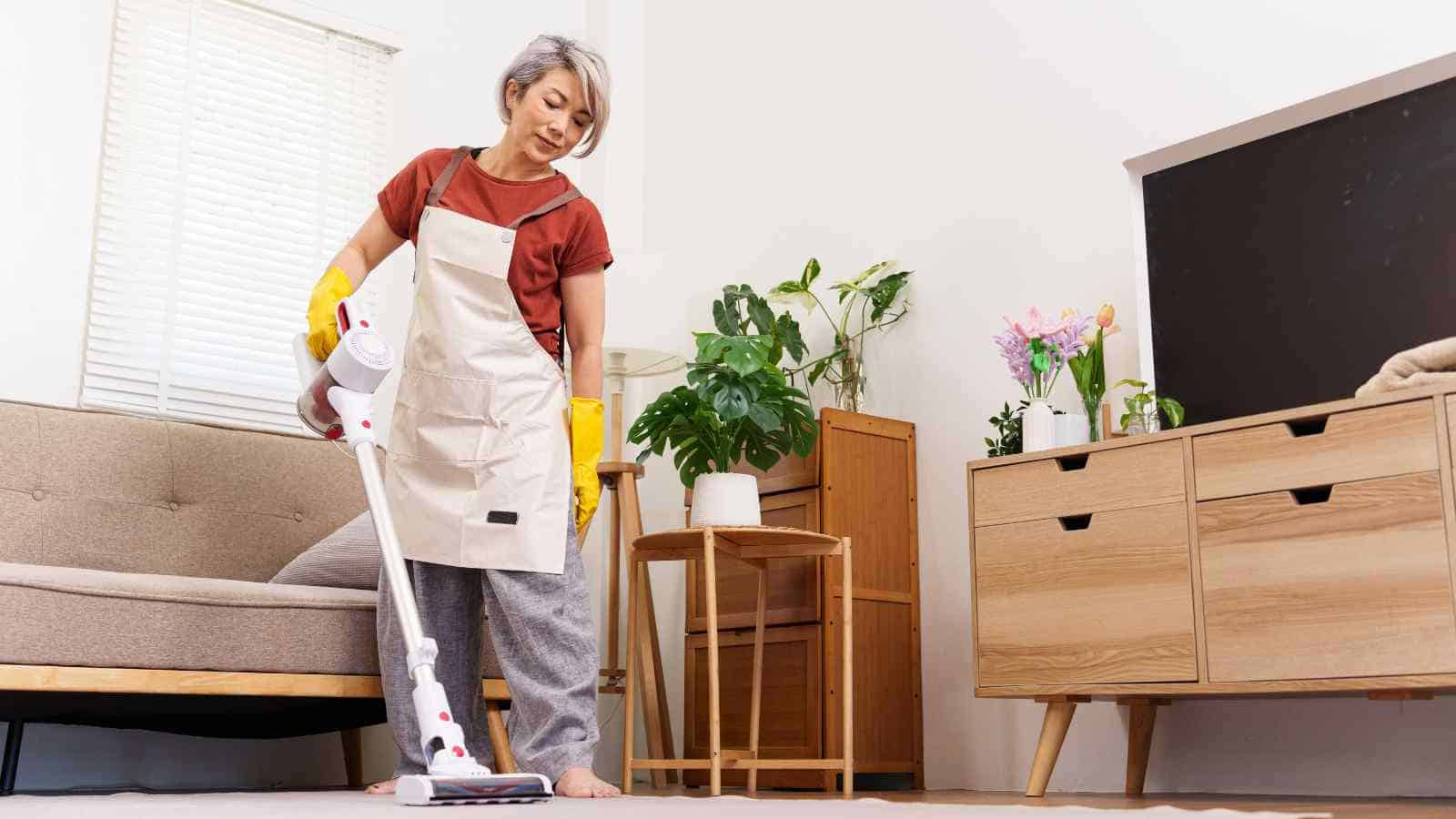 Person wearing yellow gloves and an apron vacuuming the floor in a living room with plants, a sofa, and wooden furniture.