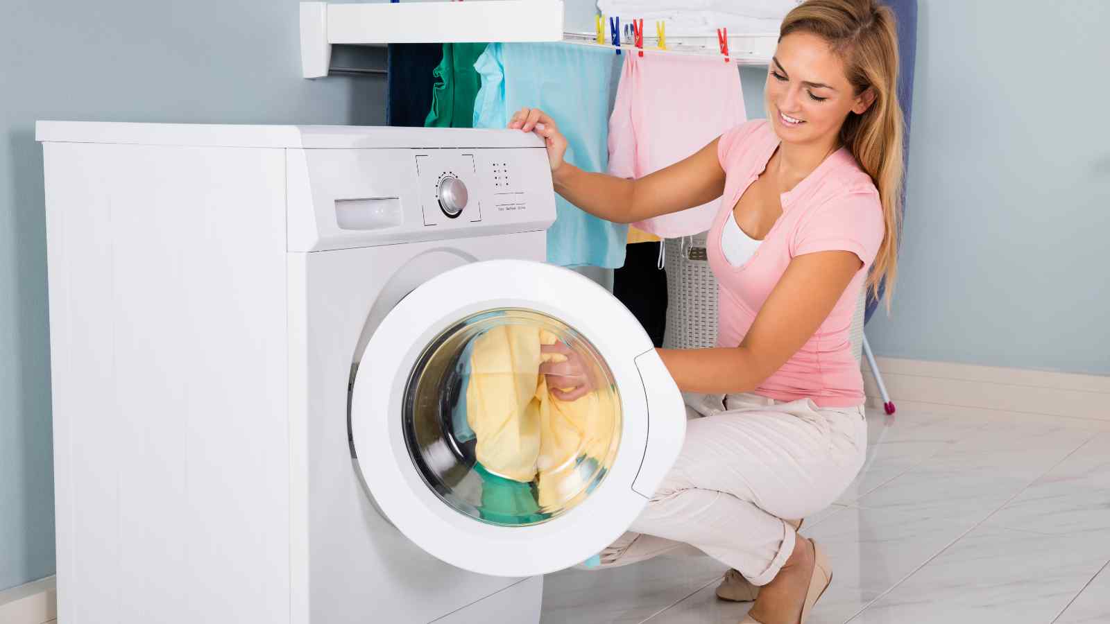 A woman kneels on the floor, loading yellow and orange laundry into a front-loading washing machine, with clothes hanging on a drying rack in the background.