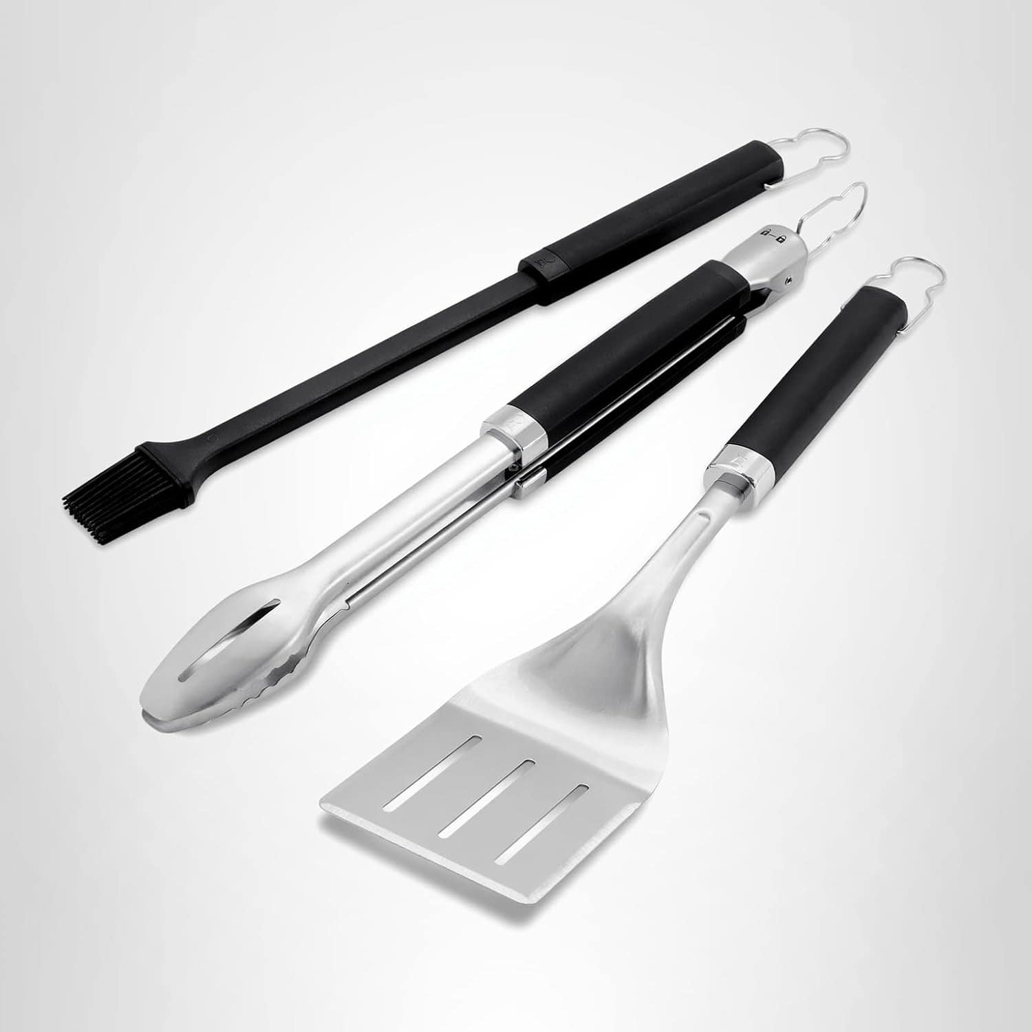 Three stainless steel barbecue tools with black handles: a basting brush, tongs, and a slotted spatula, arranged on a plain white background.