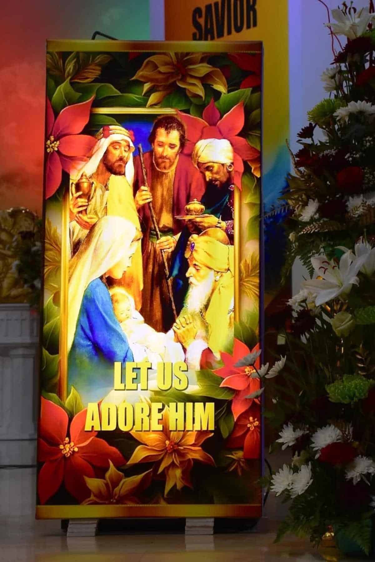 A brightly colored display shows the nativity scene with Mary, Joseph, baby Jesus, three wise men, and the text "Let Us Adore Him," surrounded by flowers.