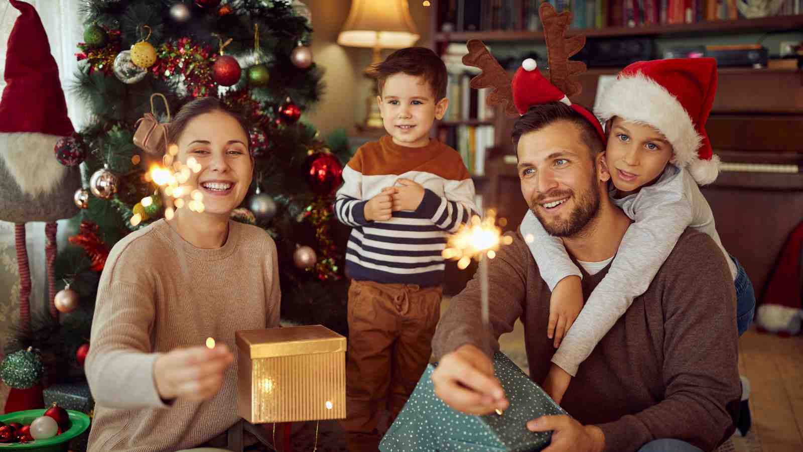 A family of four sits by a decorated Christmas tree, holding sparklers and gifts, with two children wearing festive hats.