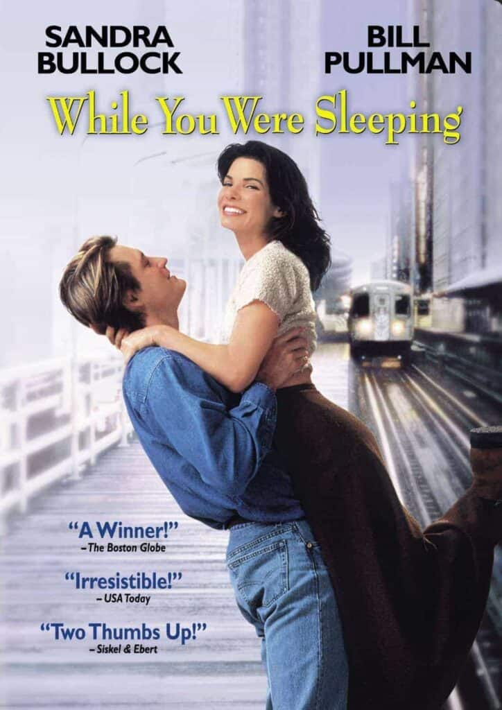 A man lifts a smiling woman in his arms on a city train platform, with the title "While You Were Sleeping" and actors' names above them.