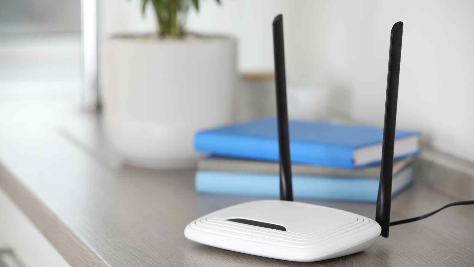 A white wireless router with two black antennas is placed on a countertop next to a potted plant and two stacked blue books.