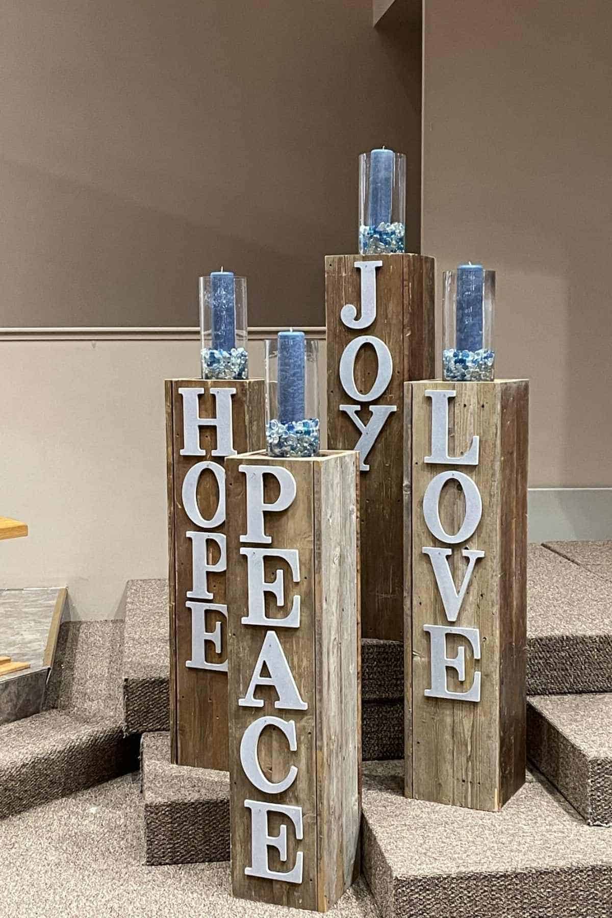 Four wooden pillars with blue candles display the words HOPE, PEACE, JOY, and LOVE in large white letters, set on a carpeted platform.