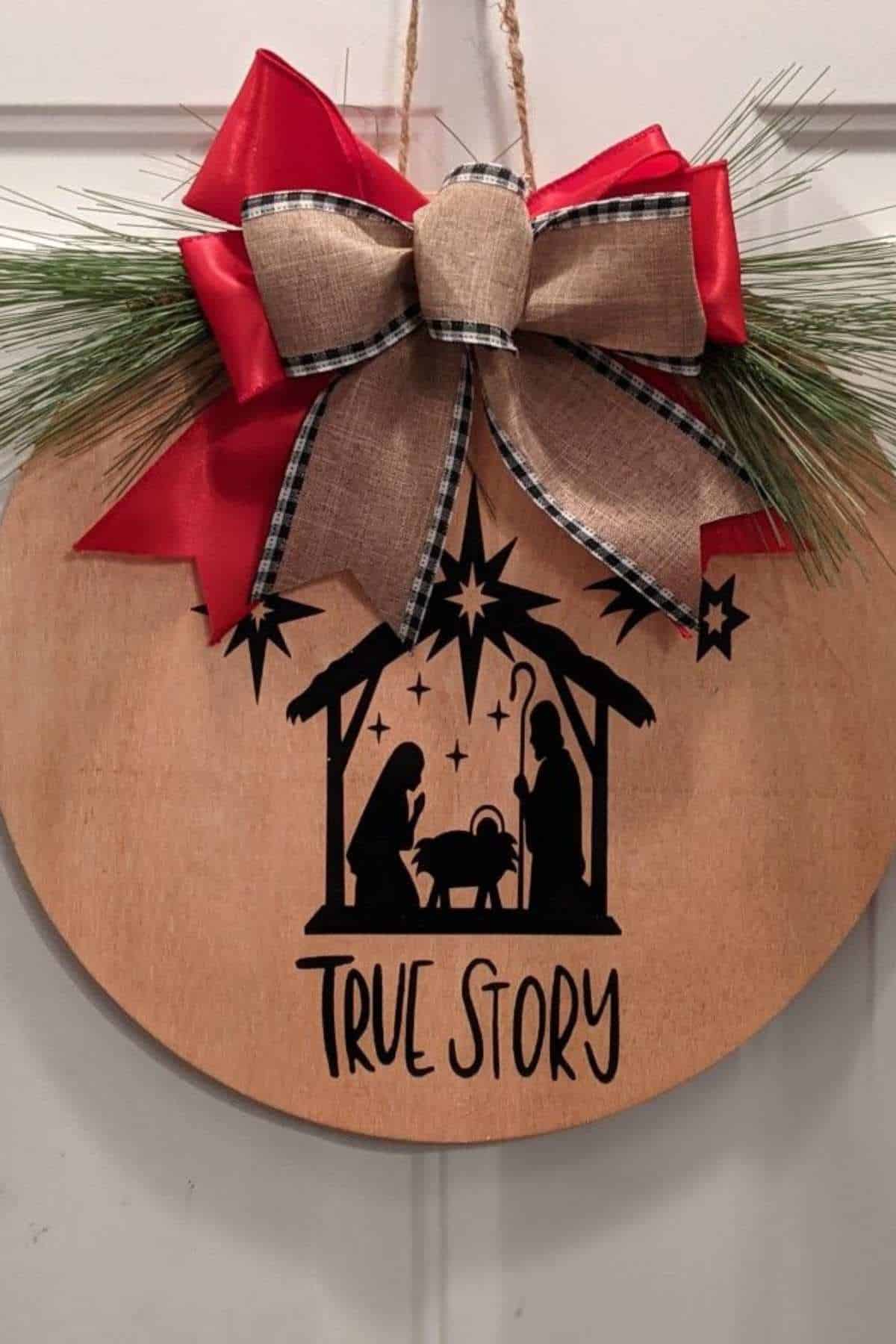 A round wooden ornament with a bow, pine needles, and a black nativity scene silhouette above the words "TRUE STORY.