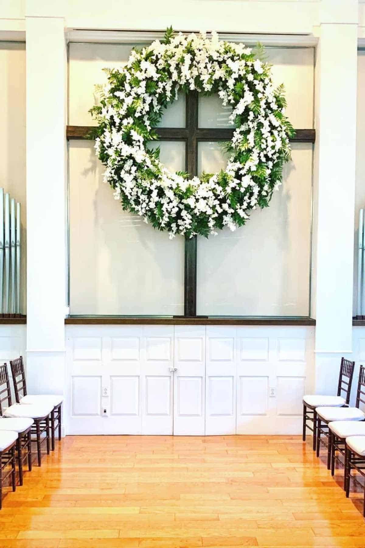 Large floral wreath with white flowers and greenery hangs on a dark wooden cross at the front of a bright room with white paneled walls and wooden chairs arranged in rows.
