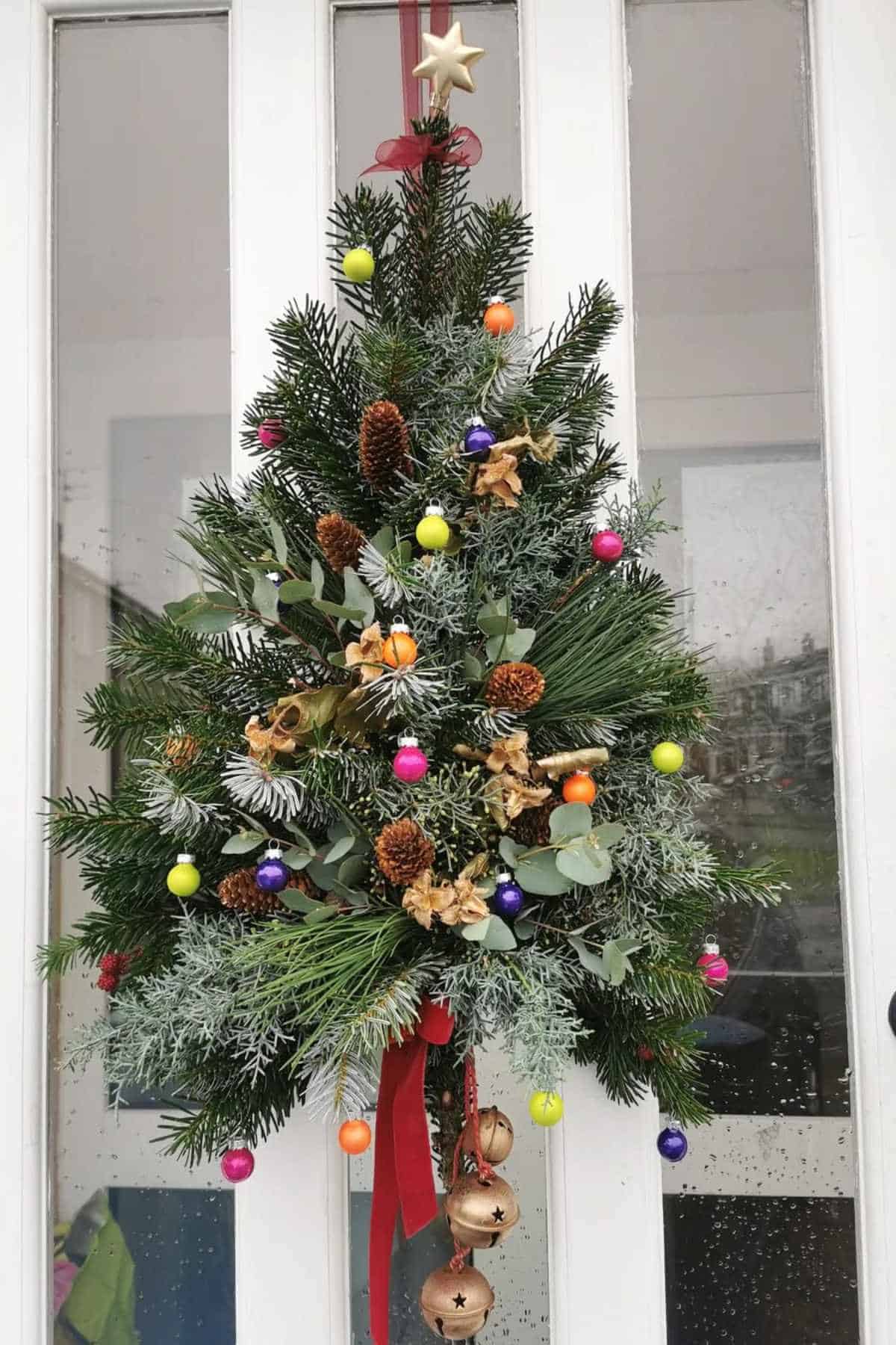 A Christmas tree-shaped wreath decorated with pinecones, dried oranges, and colorful baubles hangs on a glass door.