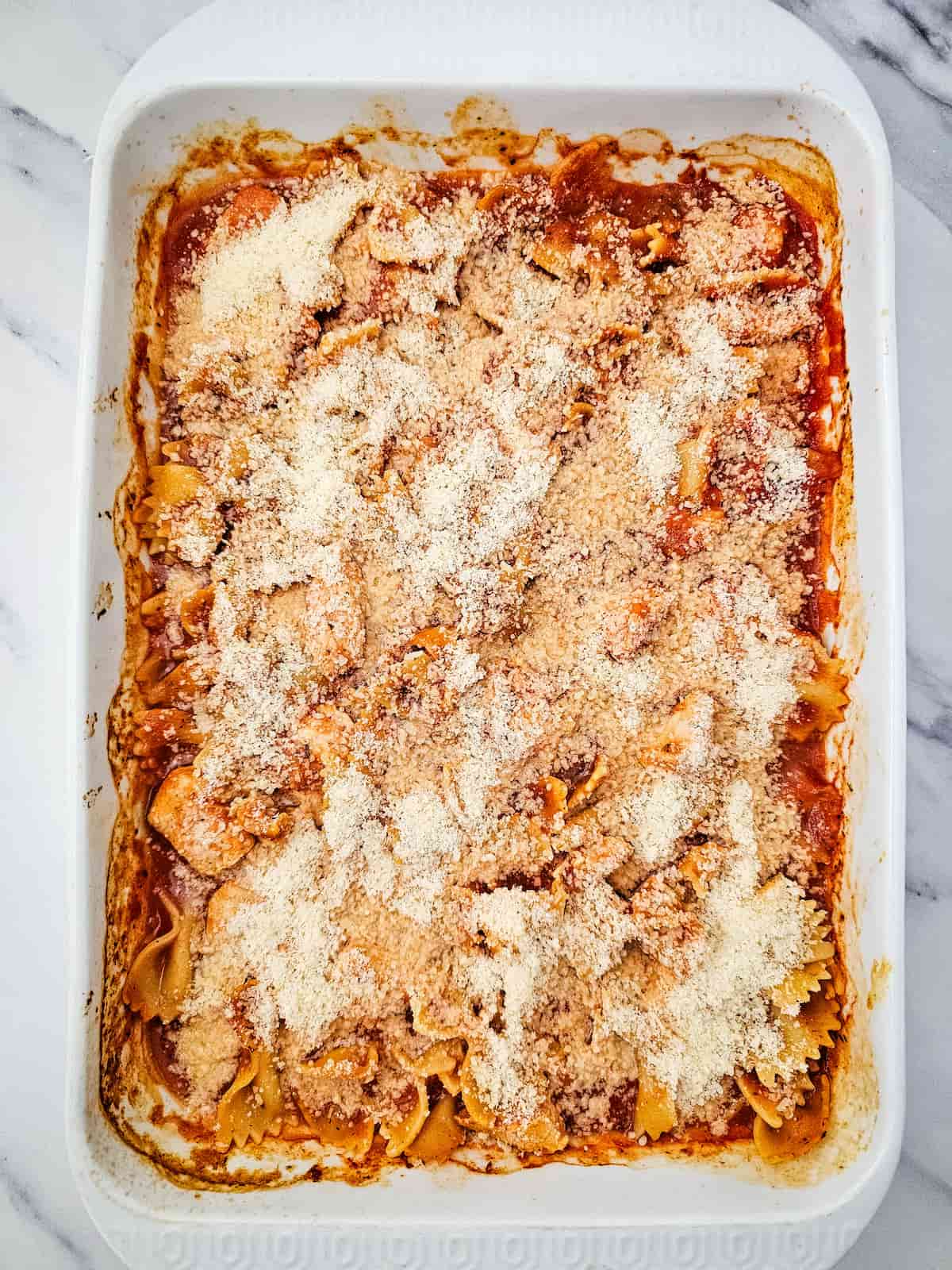 A rectangular baking dish filled with baked pasta in marinara sauce, topped with grated cheese, on a marble surface.