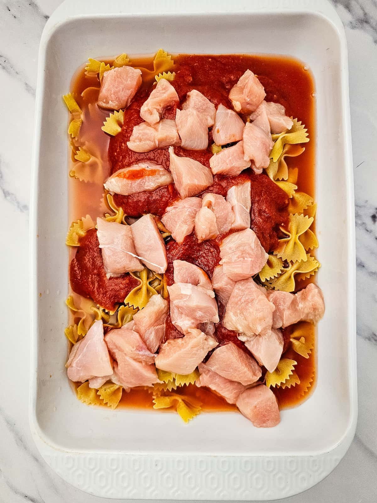 Uncooked bowtie pasta, sauce, and raw diced chicken pieces in a white rectangular baking dish on a marble surface.