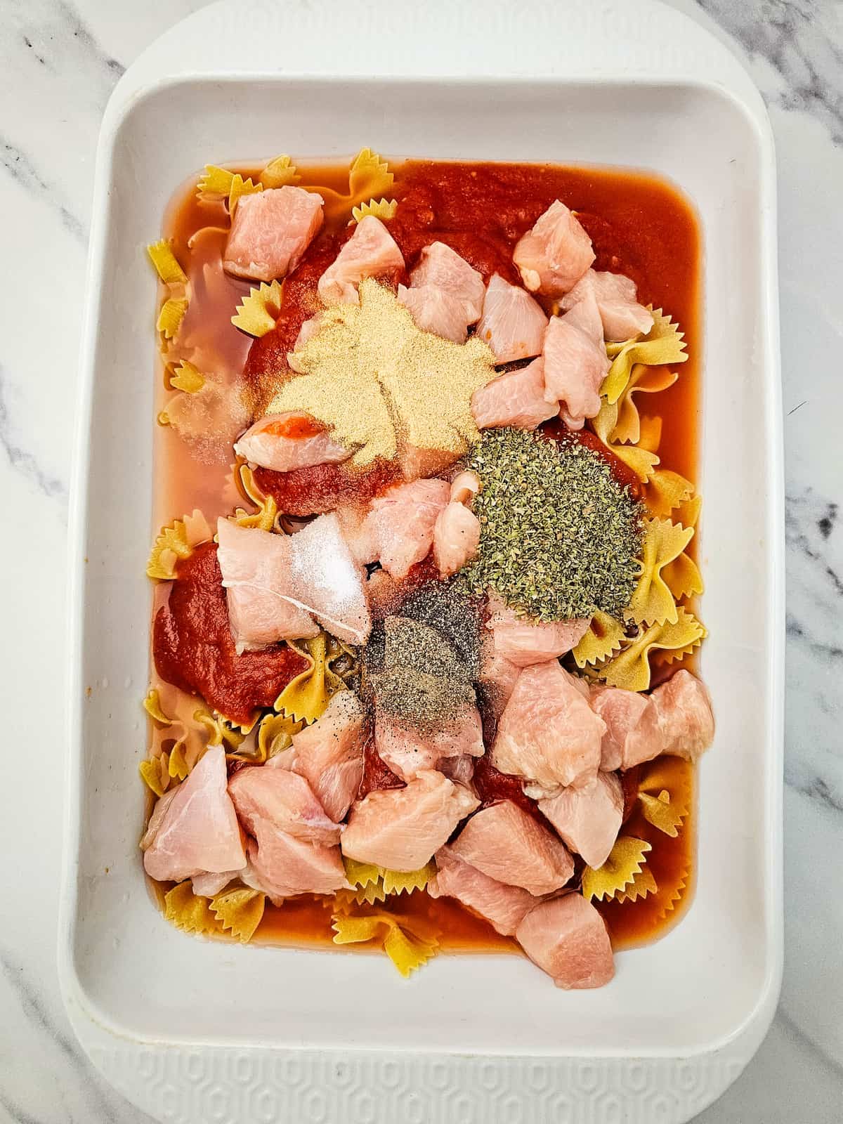 Uncooked bowtie pasta, raw chicken cubes, marinara sauce, and various seasonings are arranged in a white baking dish on a marble surface.