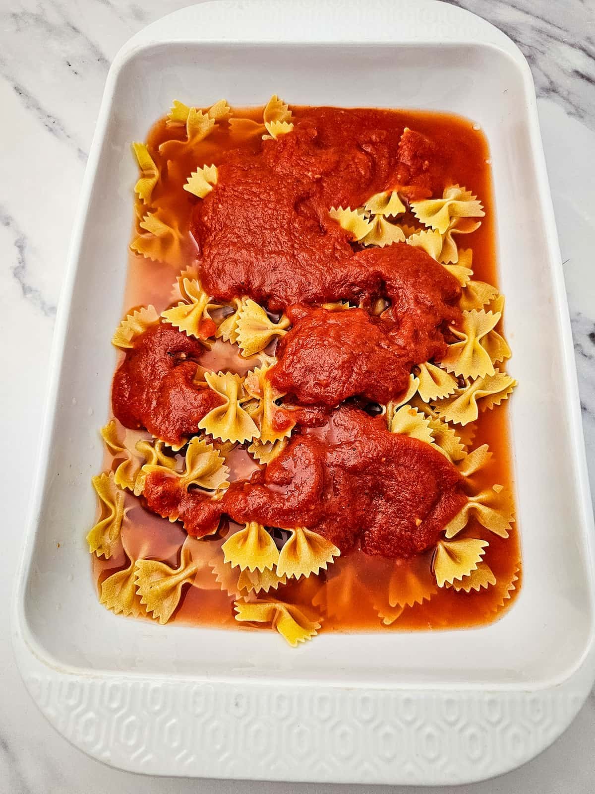 Uncooked bowtie pasta in a white baking dish topped with sauce and water on a marble surface.