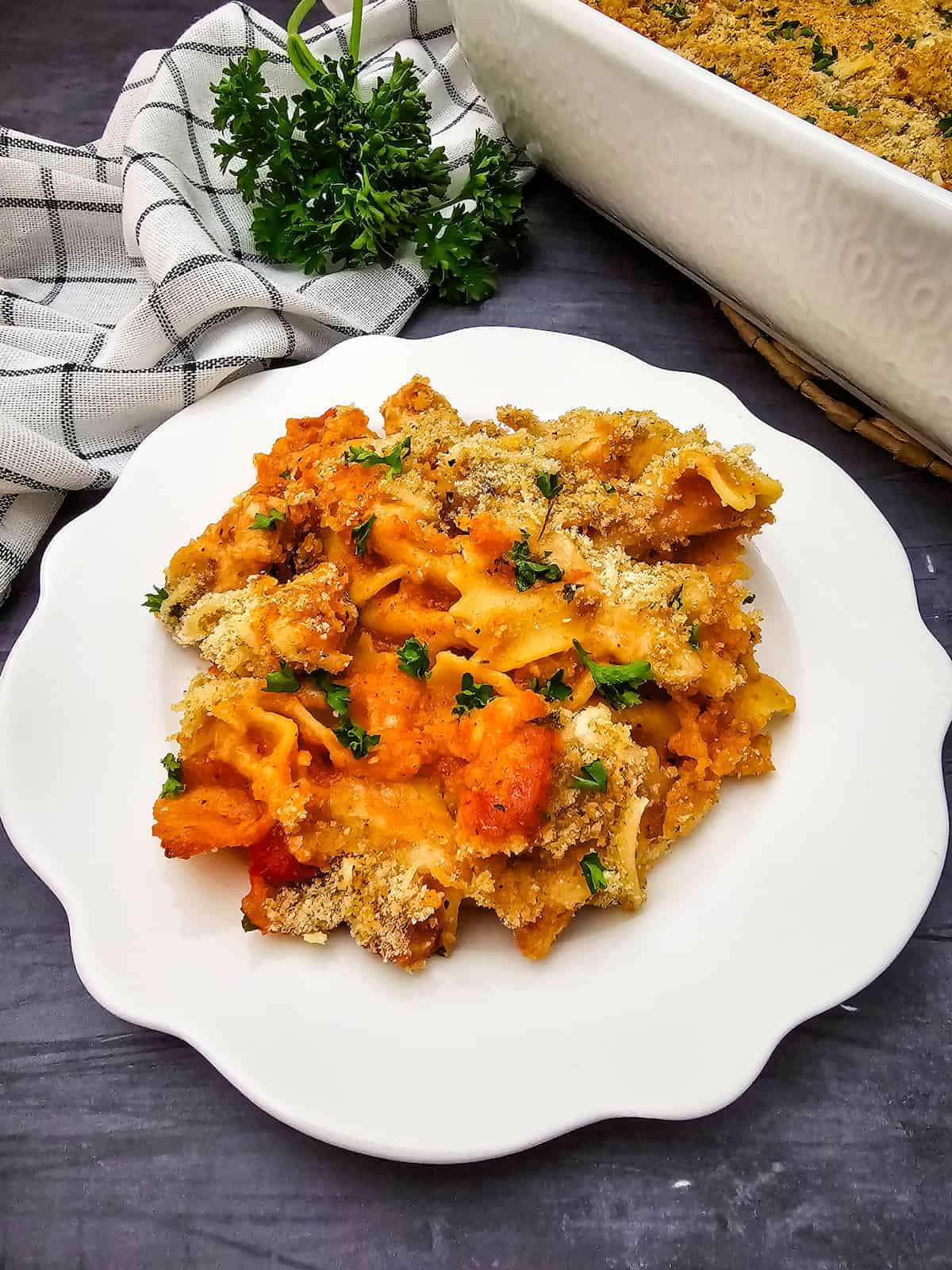 A serving of baked pasta with breadcrumbs on a white plate, garnished with parsley; a baking dish and towel are in the background.