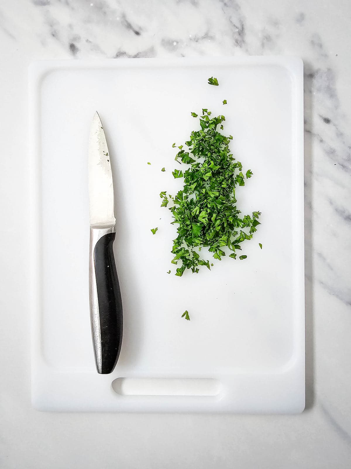 A paring knife and a small pile of chopped parsley are on a white cutting board, placed on a marble countertop.