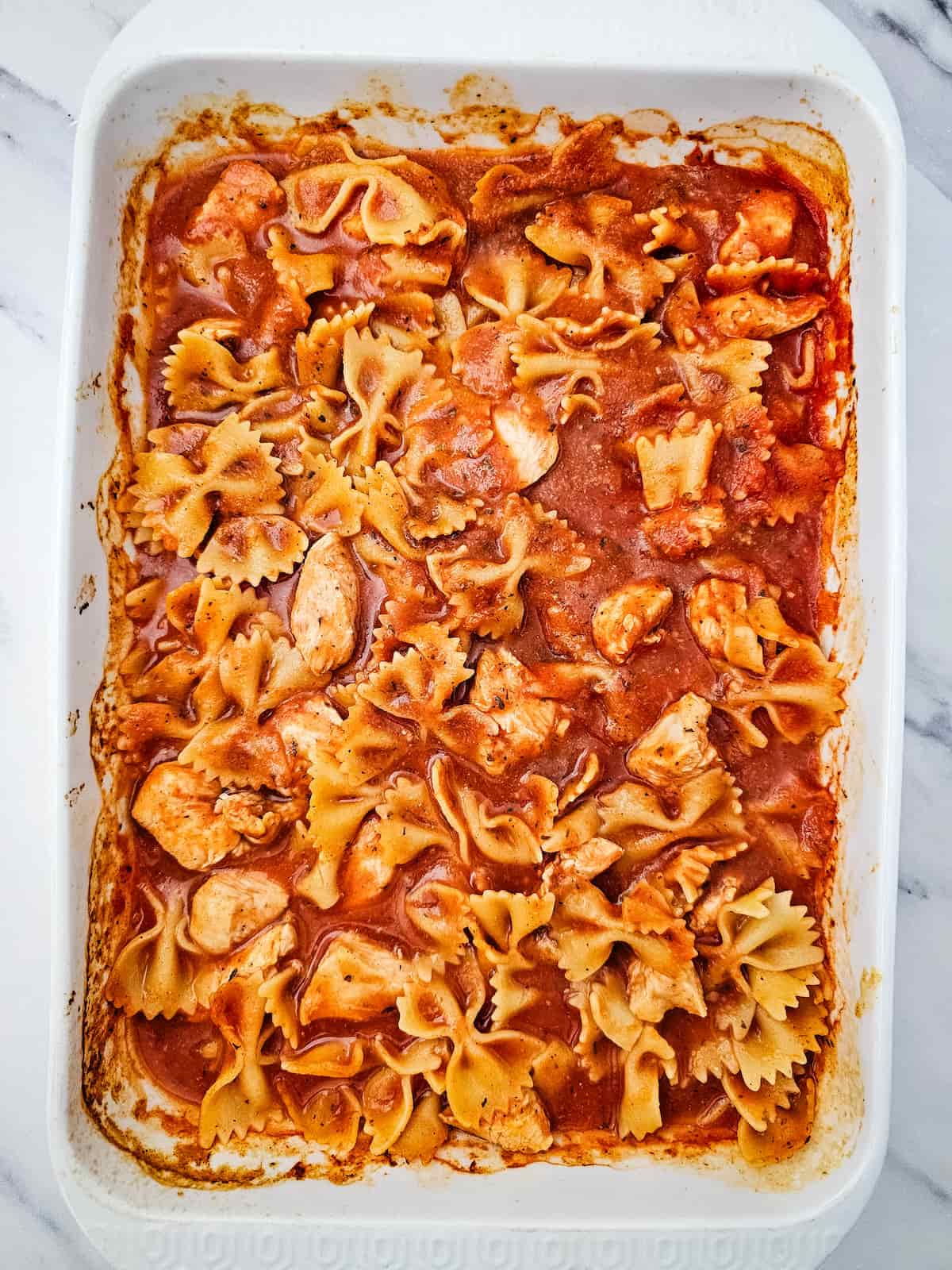 Rectangular baking dish filled with bowtie pasta, chunks of chicken, and marinara sauce on a marble surface.