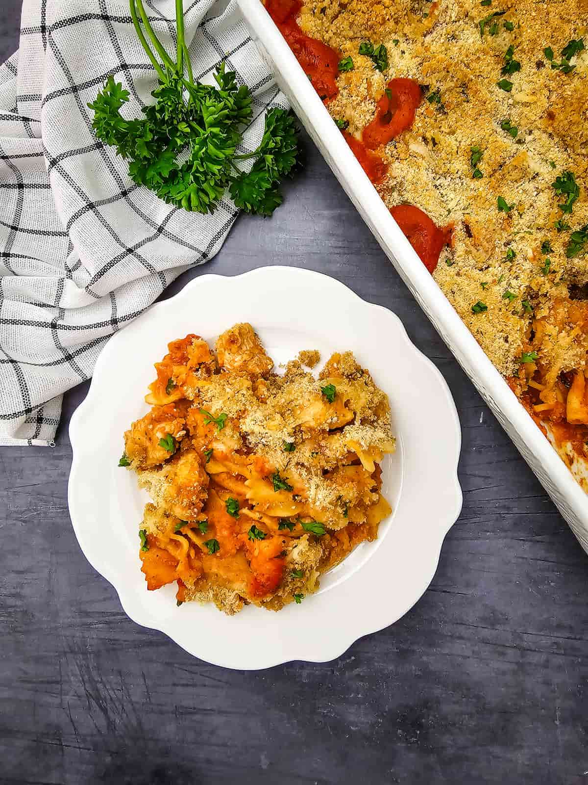 A serving of baked casserole with breadcrumb topping on a white plate, next to a baking dish and a bunch of parsley on a checkered towel.