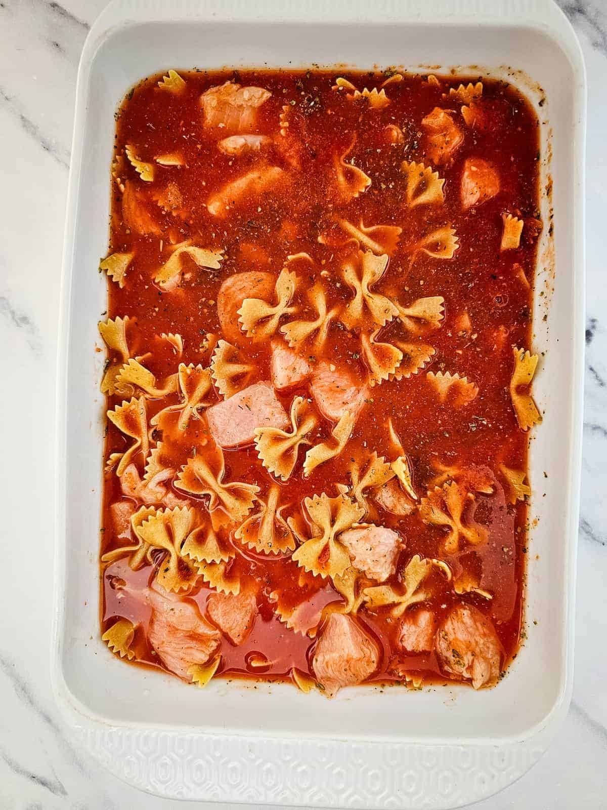 A white baking dish filled with uncooked bowtie pasta, chunks of chicken, and marinara-based sauce on a marble surface.