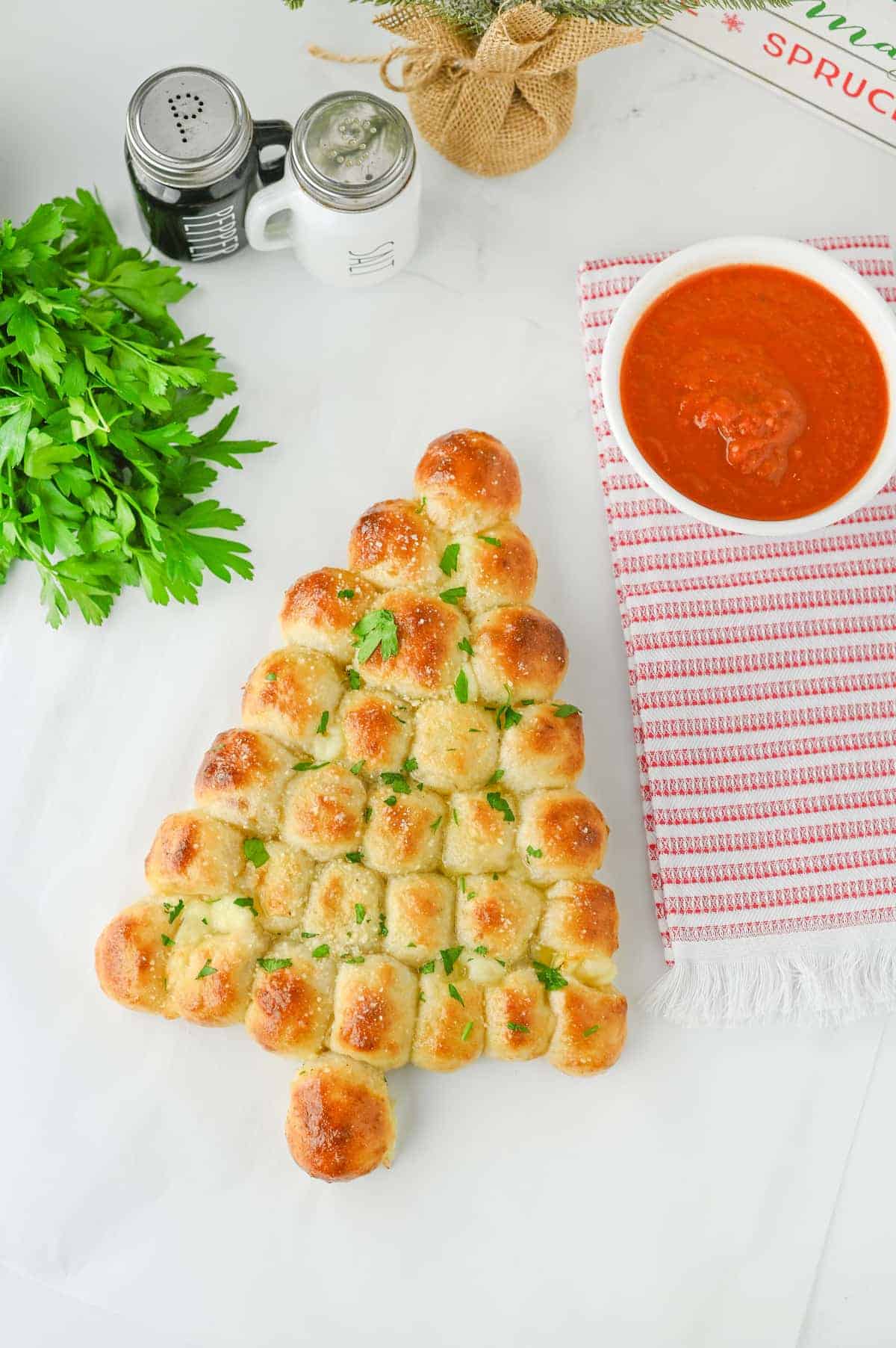 Pull-apart bread shaped like a Christmas tree, garnished with parsley, next to marinara sauce, fresh parsley, and salt and pepper shakers on a white surface.