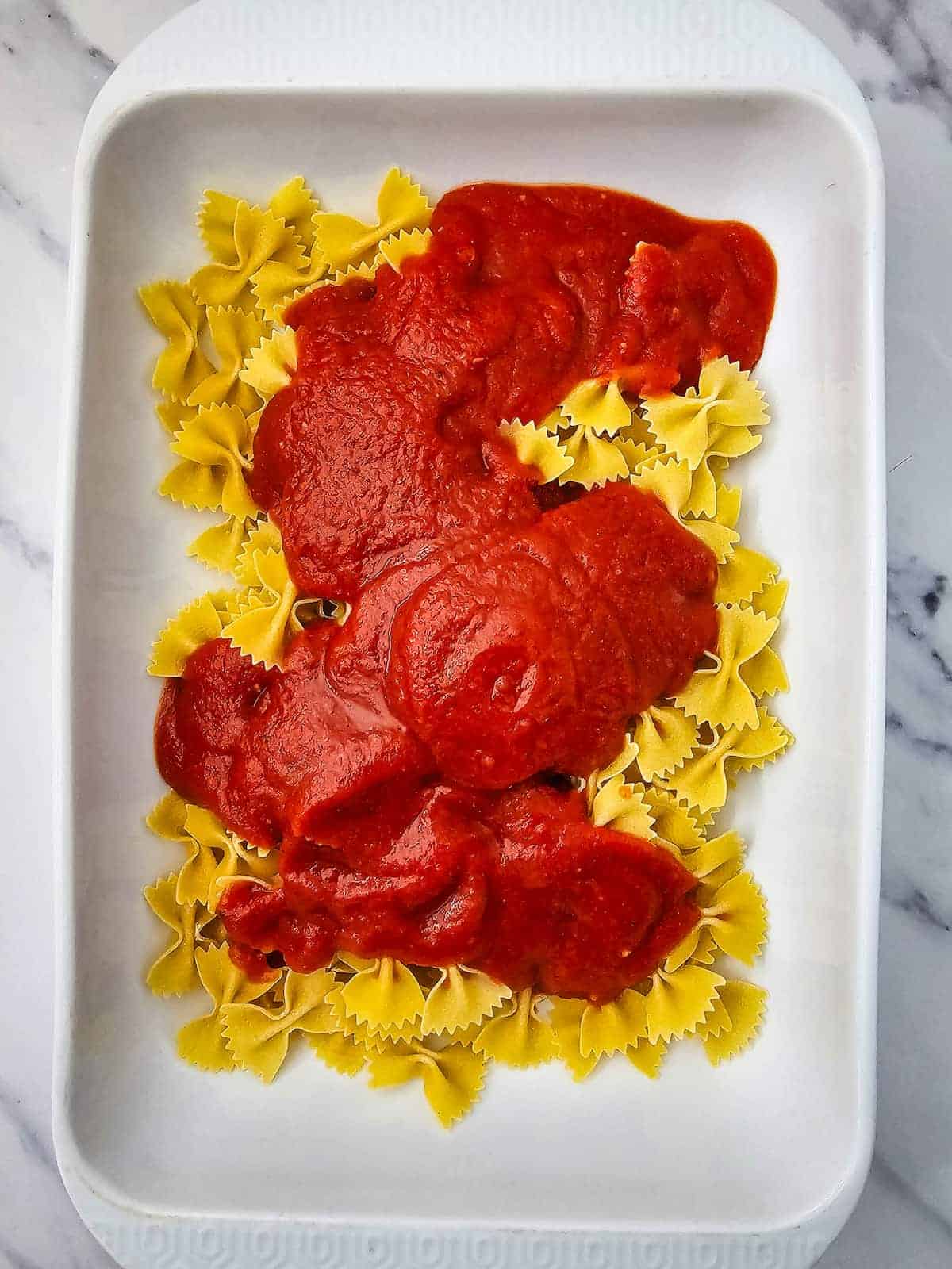 Uncooked bowtie pasta in a white baking dish topped with a generous layer of marinara sauce.