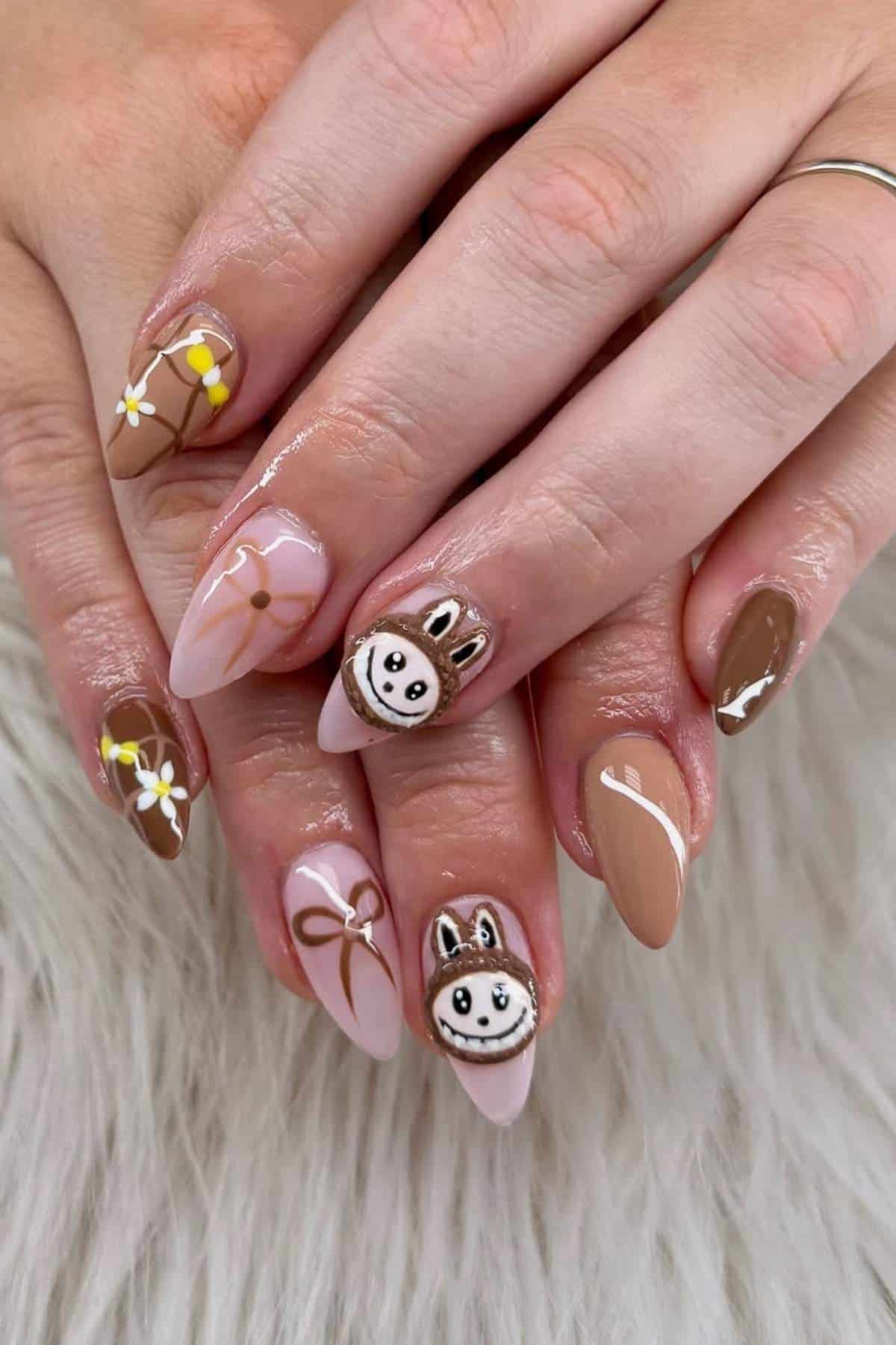 Hands with almond-shaped nails painted in brown and nude tones, featuring daisy flowers, abstract lines, and cartoon bunny faces as nail art designs.
