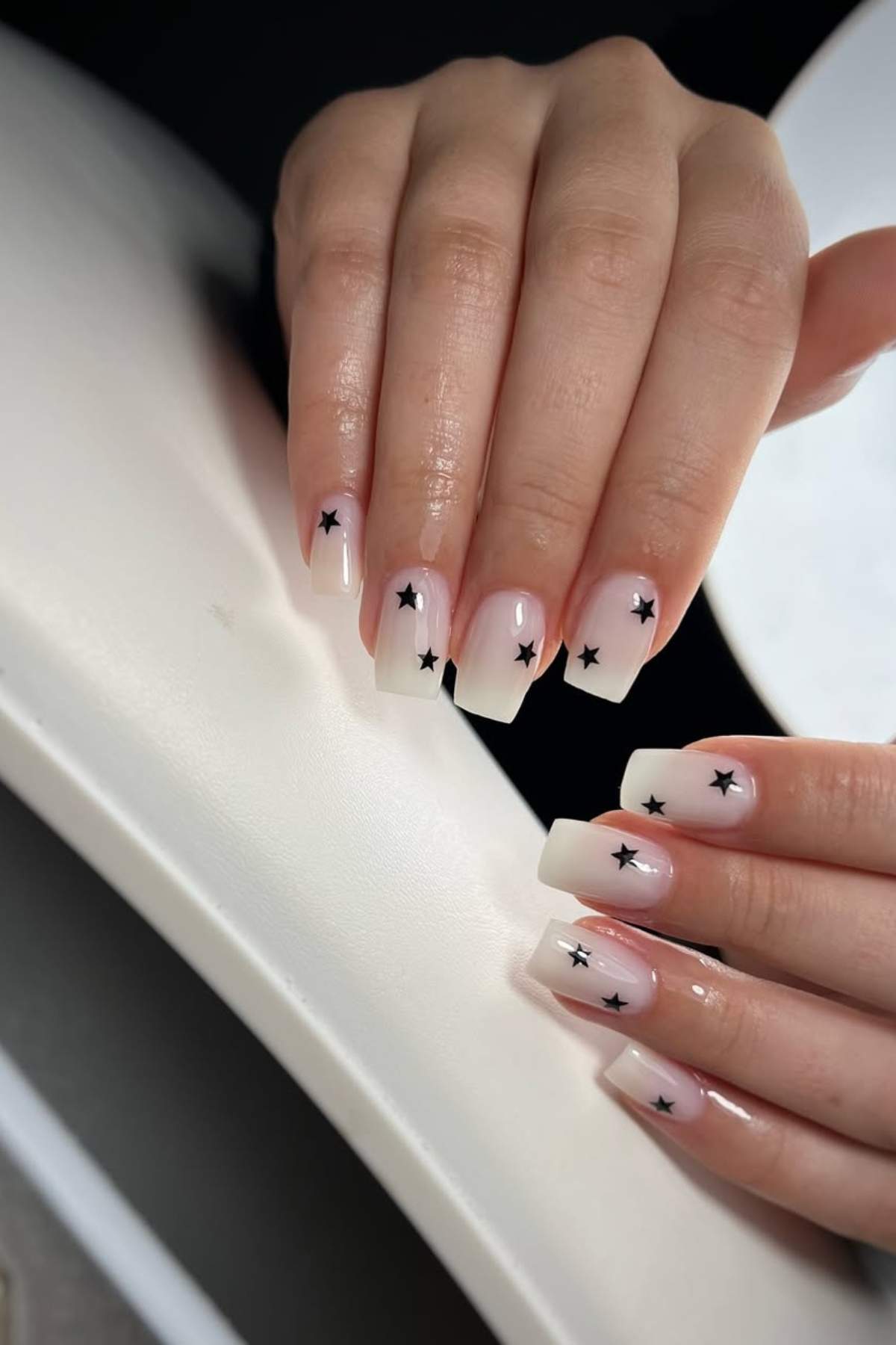 Hands with long, square-shaped nails painted in a light nude color with small black star designs on each nail.