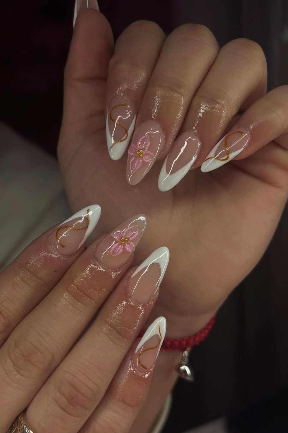 Close-up of hands with long, almond-shaped nails featuring white French tips, pink flowers, and gold accents over a clear base.